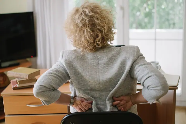 Starija žena sjedi za radnim stolom, okrenuta leđima, i drži se rukama za donji dio leđa