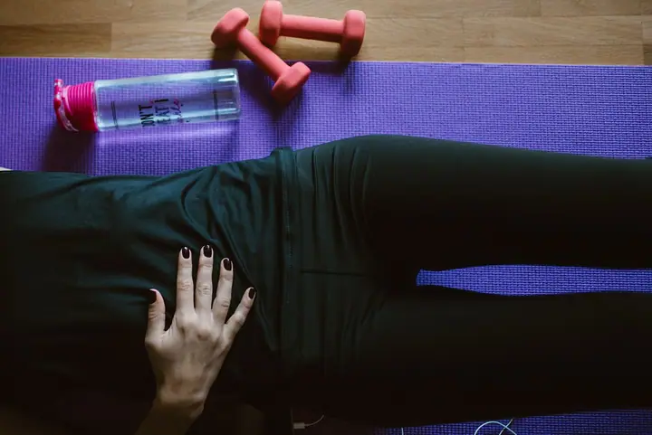 Žena u trenerci leži na ljubičastoj prostirci za vježbanje, pored nje tegovi i boca vode