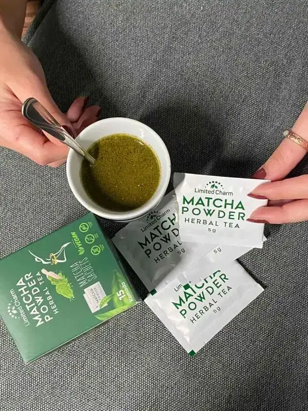 Ruke miješaju zeleni napitak u šolji, pored kesica i kutije Matcha Powder